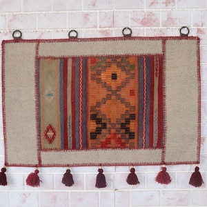 Pode incluir: Uma tapeçaria retangular com um painel central de padrões geométricos em laranja, vermelho e azul, bordada com tecido bege. A peça é finalizada com costuras em vermelho escuro, quatro argolas metálicas para pendurar e sete borlas vermelhas.