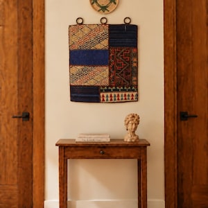 Op de afbeelding: Een rechthoekig wandkleed met een patchwork van geweven stof in blauw, rood en beige met geometrische patronen, opgehangen aan drie donkere metalen ringen. Daarboven bevindt zich een klein, rond, gevlochten mandje met groene accenten.