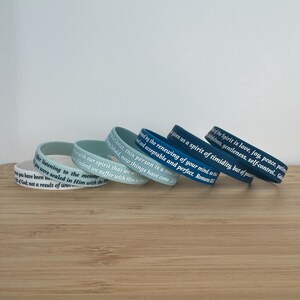 May include: A set of six silicone wristbands with different shades of blue and green. Each band features a white inscription with a bible verse.