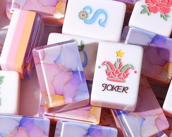 Könnte beinhalten: Eine Sammlung rechteckiger Mahjong-Spielsteine, bestehend aus weißen Steinen mit floralen und Joker-Gravuren sowie Steinen mit einem wirbelnden violetten, rosafarbenen und orangefarbenen Alkoholtintenmuster. Der Joker-Stein zeigt einen Stern und Text.