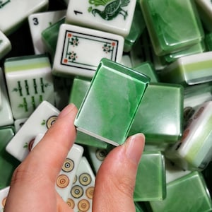 Pode incluir: Uma coleção de peças retangulares de mahjong com um acabamento marmorizado verde e branco. As peças exibem vários símbolos gravados, incluindo bambu, círculos e um pavão, com uma peça mostrando claramente o texto BAM na superfície.