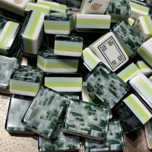 May include: A collection of rectangular mahjong tiles featuring a mix of designs. Some tiles display a tri-color pattern of white, light green, and dark green, while others show a dark green marbled texture or a white face with floral motifs.