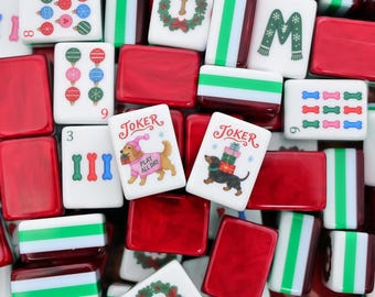 May include: A collection of mahjong tiles featuring Christmas-themed illustrations including colorful ornaments, wreaths, dog bones, and scarves. Two tiles are labeled Joker, one showing a dog in a pink sweater and one with a stack of gifts.