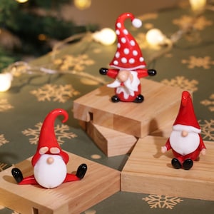 Może przedstawiać: Trzy miniaturowe świąteczne gnomy z czerwonymi czapkami i białymi brodami. Jeden gnom siedzi, drugi stoi, a trzeci ma czapkę w kropki. Są one wystawione na małych drewnianych klockach z świątecznym tłem.