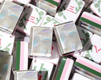 Könnte beinhalten: Eine Sammlung von Mahjong-Spielsteinen, bestehend aus weißen Steinen mit roten und grünen Blumen- oder Bambusgravuren sowie Steinen mit perlmuttartiger, schillernder Oberfläche. Einige Steine zeigen ein Muster aus rosa, weißen und dunkelgrünen Streifen.