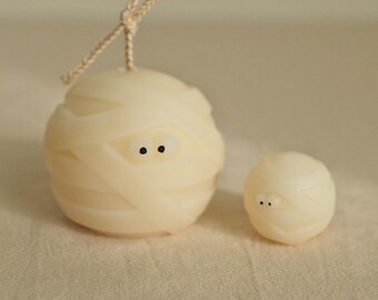 May include: Two white mummy-shaped candles with black eyes. The larger candle has a string attached to the top.