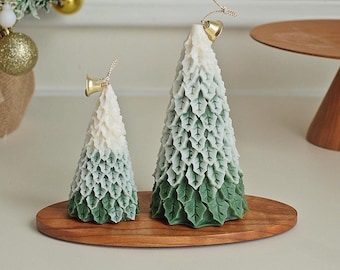 May include: Two green and white ombre Christmas tree candles with gold bells on top, sitting on a wooden tray.