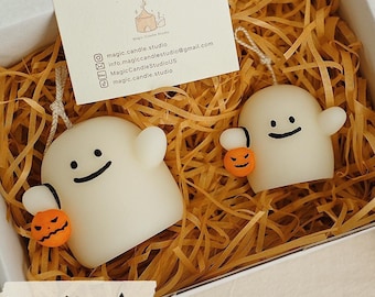 May include: Two white ghost-shaped candles with black eyes and mouths, each holding a small orange pumpkin candle. The candles are in a white box filled with shredded brown paper. The box is sitting on a table with a white pumpkin and an orange pumpkin.