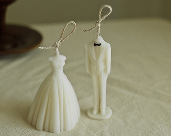 May include: Two white wedding cake toppers, one in the shape of a dress and the other in the shape of a tuxedo, with a bow tie. The toppers are on a white surface with three small pink and purple candles that spell "I Love You".