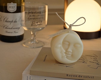 May include: A white candle shaped like a crescent moon with two faces on it. The candle is sitting on top of two books. The top book is brown and has the text "Moments Precious" on the spine. The bottom book is white and has the text "Matisse Cut-out" on the front cover. The text "Rayon de soleil" is printed on the brown book.