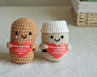 May include: Two crocheted plushies, one shaped like a coffee cup and the other like a bean, holding heart-shaped cards with messages. The coffee cup plush is brown and white, and the bean plush is brown. The cards are red with white text. The coffee cup plush says "I can't espresso how much you mean to me!" and the bean plush says "You are my favorite human Bean!"