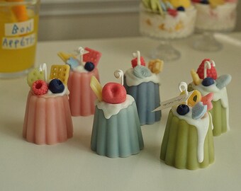 May include: Assortment of six small, colorful, decorated candles shaped like individual desserts. Each candle has a different topping, including fruit, sprinkles, and cookies. The candles are arranged on a white surface.