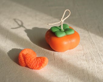 May include: Two orange persimmon shaped candles, one with a green leaf detail and a string tied around the top.