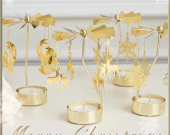 May include: Three gold metal candle holders with a spinning carousel design. Each holder features a different Christmas design, including a reindeer, a snowflake, and a Christmas tree. The candle holders are on a white surface with the text "Merry Christmas" in the foreground.