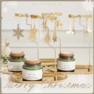 May include: A festive Christmas candle set with three glass jars of green scented candles, each with a cork lid. Above, a gold-colored carousel with decorative ornaments, including snowflakes, trees, and reindeer, spins above the candles. The words "Merry Christmas" are visible.