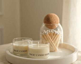 May include: Two white candles in glass jars with the text "Magic Candle" on the front. A glass jar with a cork lid filled with wooden matches is on a white round tray.