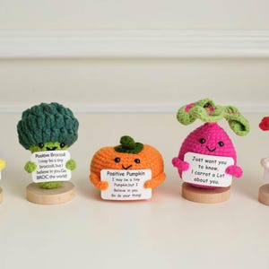 May include: A collection of five crocheted vegetable and fruit figures, including an apple, broccoli, pumpkin, radish, and mushroom. Each figure is brightly colored and holds a small sign with a positive message. They are displayed on small wooden bases.