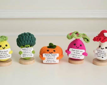 May include: A collection of five crocheted vegetable and fruit figures, including an apple, broccoli, pumpkin, radish, and mushroom. Each figure is brightly colored and holds a small sign with a positive message. They are displayed on small wooden bases.