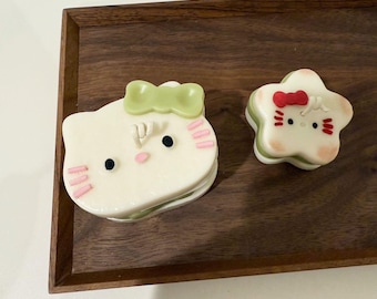 May include: Two small, white, layered cakes decorated with pink and green frosting. One cake is shaped like a cat with a green bow, and the other is shaped like a star with a red bow. Both cakes have black eyes and pink cheeks.