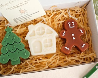 May include: A set of three Christmas-themed candles in a white box with shredded paper. The candles are shaped like a gingerbread man, a house, and a Christmas tree. The gingerbread man is brown with white icing details, the house is white with a brown door and windows, and the Christmas tree is green with white dots.