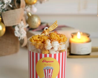 May include: A red and white striped glass jar filled with popcorn-shaped candles. The jar has a yellow label with the word "Popcorn" and a cartoon image of a red and white striped popcorn bucket.