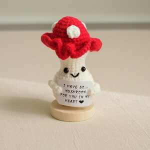 May include: A crocheted mushroom holding a sign that says "I have so... mushroom for you in my heart." The mushroom is white with a red cap and a happy face. It is standing on a wooden base.