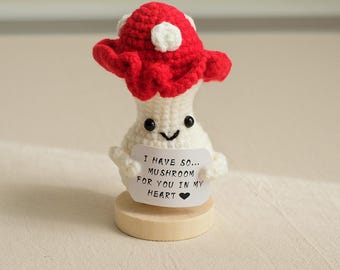 May include: A crocheted mushroom holding a sign that says "I have so... mushroom for you in my heart." The mushroom is white with a red cap and a happy face. It is standing on a wooden base.