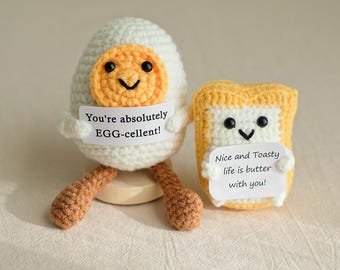 May include: Two crocheted plush toys, one shaped like an egg and the other like a slice of bread. The egg is white with a yellow yolk and has a smiling face. The bread is yellow and has a smiling face. Both toys are holding signs with messages. The egg's sign says "You're absolutely EGG-cellent!" and the bread's sign says "Nice and Toasty life is butter with you!"