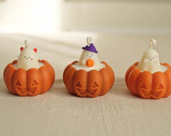 May include: Three Halloween-themed candles shaped like pumpkins with ghost figures inside. The pumpkins are orange with a jack-o'-lantern face. The ghosts are white with black eyes and mouths. One ghost is wearing a purple hat.