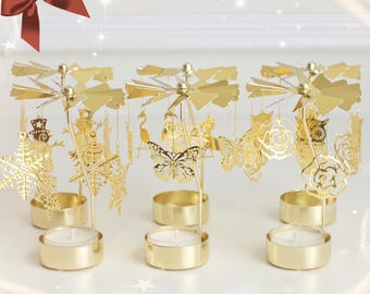 May include: Three gold metal candle holders with a carousel design. Each holder features a different set of decorative cutouts, including butterflies, snowflakes, and flowers. The candle holders are on a white surface.