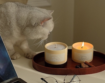 May include: Two candles on a wooden tray with a cat in the background. One candle is lit and the other is unlit. Both candles are in glass jars with a cream-colored label that says "Happy Hour Candle Co."