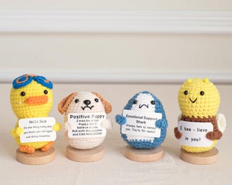 May include: Four crocheted amigurumi figures: a duck, a puppy, a shark, and a bee. Each figure holds a small sign with a positive message. The duck wears goggles, the puppy has brown ears, the shark is blue, and the bee has wings.