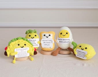 May include: A collection of five crocheted food items: a taco, apple, toast, egg, and lemon. Each item has a smiling face and a small sign with a positive message. The taco says "Emotional Support Taco". The egg says "You're absolutely EGG-cellent!"