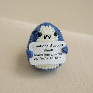 May include: A blue and white crocheted shark holding a sign that says "Emotional Support Shark Always here to remind you: You're fin-tastic!"