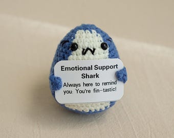 May include: A blue and white crocheted shark holding a sign that says "Emotional Support Shark Always here to remind you: You're fin-tastic!"