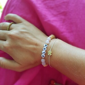 Könnte beinhalten: Eine Nahaufnahme eines Handgelenks, an dem zwei Armbänder getragen werden. Ein Armband besteht aus hellrosa und braunen Perlen, auf denen das Wort "FLOW" in weißen Buchstaben geschrieben ist. Das andere Armband ist ein dünnes weißes Band mit einem vierblättrigen Kleeblatt-Anhänger aus Gold.