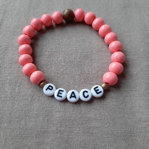 Könnte beinhalten: Ein rosa Perlenarmband mit einer braunen Holzperle und weißen Buchstabenperlen, die das Wort "FRIEDEN" buchstabieren.
