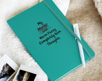 May include: A teal blue notebook with the text "My Murder List Warm Fuzzy Completely Sane Thoughts" written on the front. The notebook is lying on a white and brown fuzzy surface with a pen and a pair of white headphones.