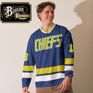 Op de afbeelding: Een blauw hockeytrui met lange mouwen met gele en witte strepen. Het trui heeft het woord "CHIEFS" in gele letters op de voorkant.