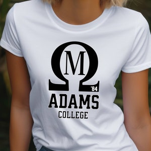 Könnte beinhalten: Weißes T-Shirt mit schwarzem Aufdruck, der den griechischen Buchstaben Omega, den Buchstaben M und die Worte "ADAMS COLLEGE" mit "'84" darunter zeigt. Das Shirt stammt von Boxcar Bandit Apparel.