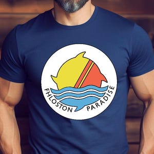 Op de afbeelding: Marineblauw T-shirt met een cirkelvormige afbeelding. De afbeelding toont een gele en rode dolfijn die over blauwe golven springt, met de woorden "FHLOSSTON PARADISE" in het wit. Het T-shirt is gemaakt van zacht materiaal.