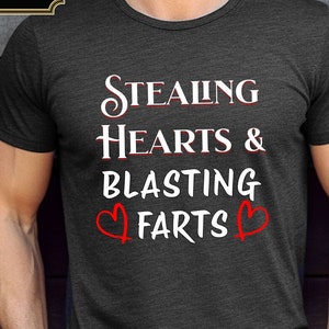 May include: A dark grey t-shirt with the text "STEALING HEARTS & BLASTING FARTS" in white with two red hearts on either side of the word "FARTS".