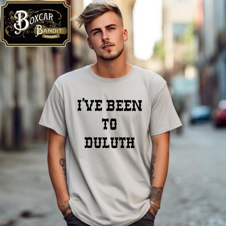 May include: A light gray t-shirt with the text "I'VE BEEN TO DULUTH" in black, block letters. The shirt also features a logo in the upper left corner. The t-shirt is a casual apparel item.