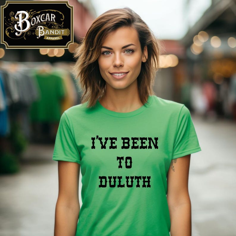 May include: A green t-shirt with the text "I'VE BEEN TO DULUTH" in black, block letters. The shirt is worn by a person. The Boxcar Bandit Apparel logo is in the upper left corner.