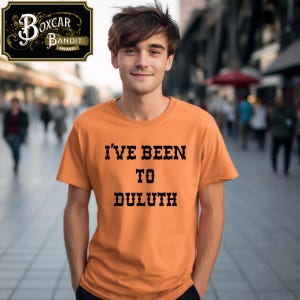 May include: Orange t-shirt with the text "I'VE BEEN TO DULUTH" in black, block letters. The shirt is short-sleeved. The Boxcar Bandit Apparel logo is in the upper left corner. A person is wearing the shirt.