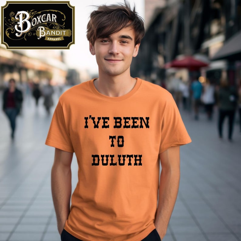 May include: Orange t-shirt with the text "I'VE BEEN TO DULUTH" in black, block letters. The shirt is short-sleeved. The Boxcar Bandit Apparel logo is in the upper left corner. A person is wearing the shirt.