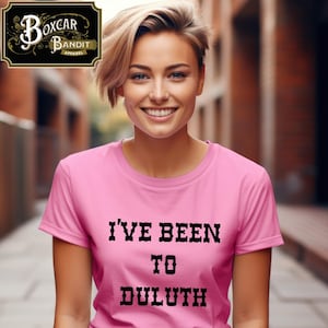 May include: A pink t-shirt with the text "I'VE BEEN TO DULUTH" in black, bold letters. The person is smiling and wearing the t-shirt. The Boxcar Bandit Apparel logo is in the upper left corner.
