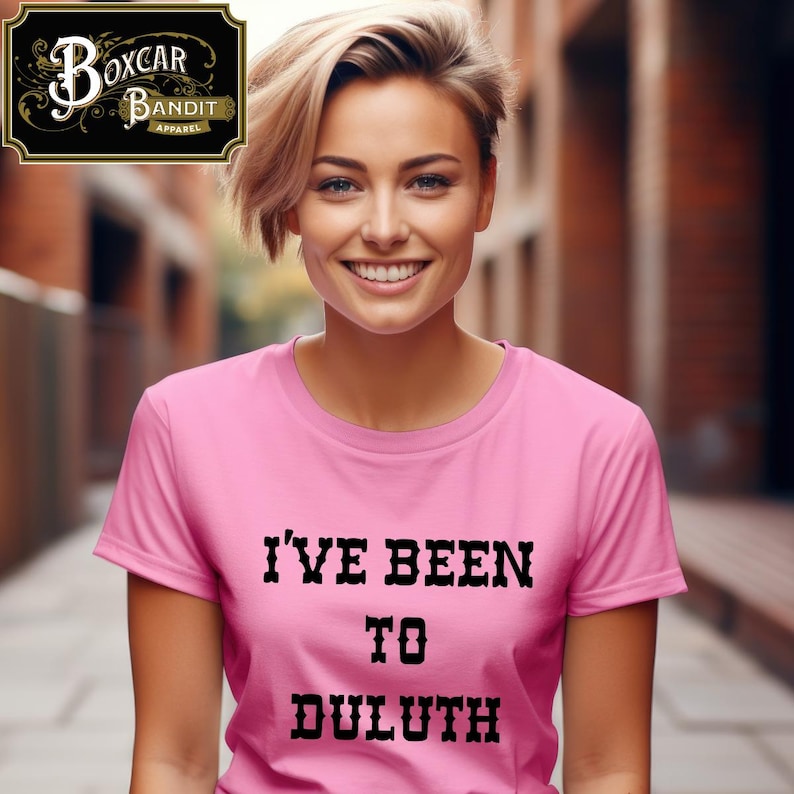 May include: A pink t-shirt with the text "I'VE BEEN TO DULUTH" in black, bold letters. The person is smiling and wearing the t-shirt. The Boxcar Bandit Apparel logo is in the upper left corner.