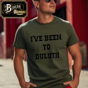 May include: Olive green t-shirt with the text "I'VE BEEN TO DULUTH" in black block letters. The shirt is a crew neck style and appears to be made of a soft material. The Boxcar Bandit Apparel logo is visible in the upper left corner.
