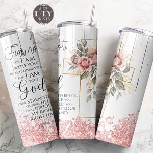 May include: A white tumbler with a pink glitter bottom and a floral design featuring a cross and pink roses. The tumbler has the text "Fear not, for I am with you; be not dismayed, for I am your God" and a Bible verse.